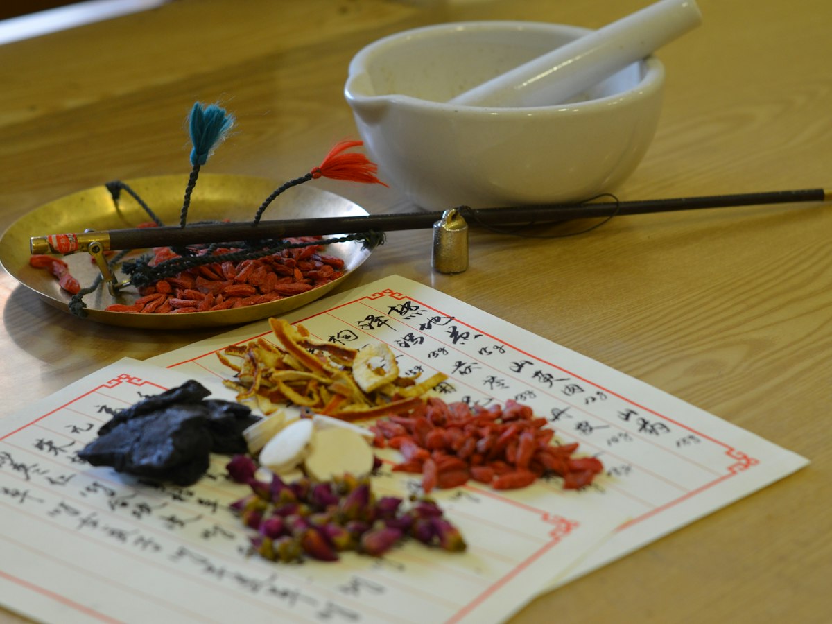 Dried herbs, goji berries and rosebuds on a handwritten Chinese prescription sheet, beside a brass hand scale and a white mortar and pestle